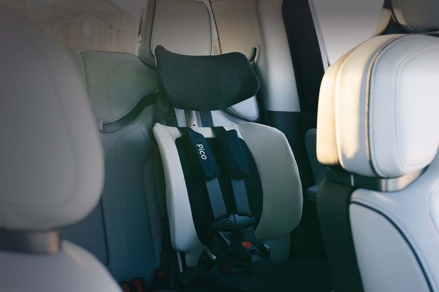 A Pico car seat installed in the back of a vehicle, shown from a side-rear angle. The car interior features light-colored leather seats, and the Pico brand name is visible on the harness strap.