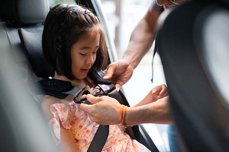 A young girl wearing a floral dress sits in a car seat while an adult fastens her car seat belt. The photo is taken inside a vehicle with a blurred gray background.