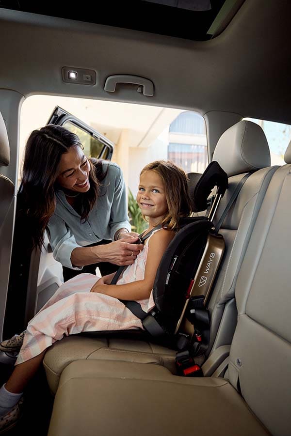 A woman in a light blue shirt is fastening a child's seatbelt in a vehicle's rear passenger area. The child is seated in a black and tan Wayb car seat.